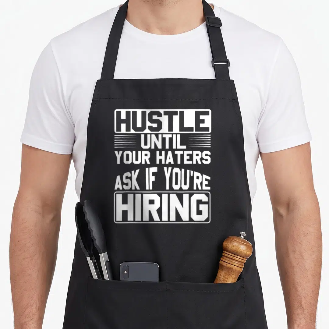 Hater hustle apron - White stacked text reading HUSTLE UNTIL YOUR HATERS ASK IF YOU'RE HIRING with graphic borders — shown on a black cotton canvas apron against a white background
