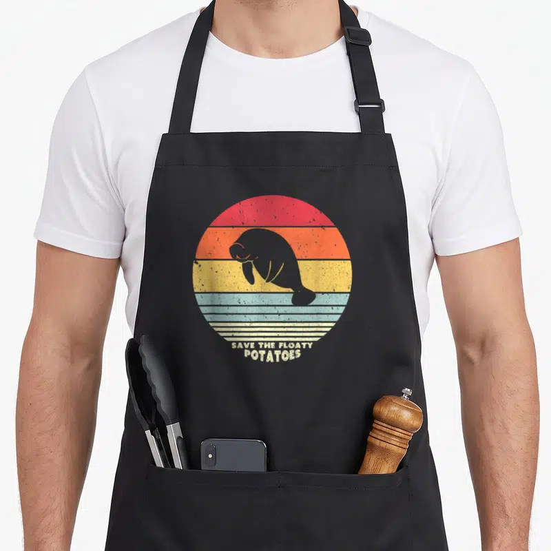 Save manatee lover apron - Grey manatee silhouette inside red orange yellow blue striped circle with text SAVE — shown on a black cotton canvas apron against a white background