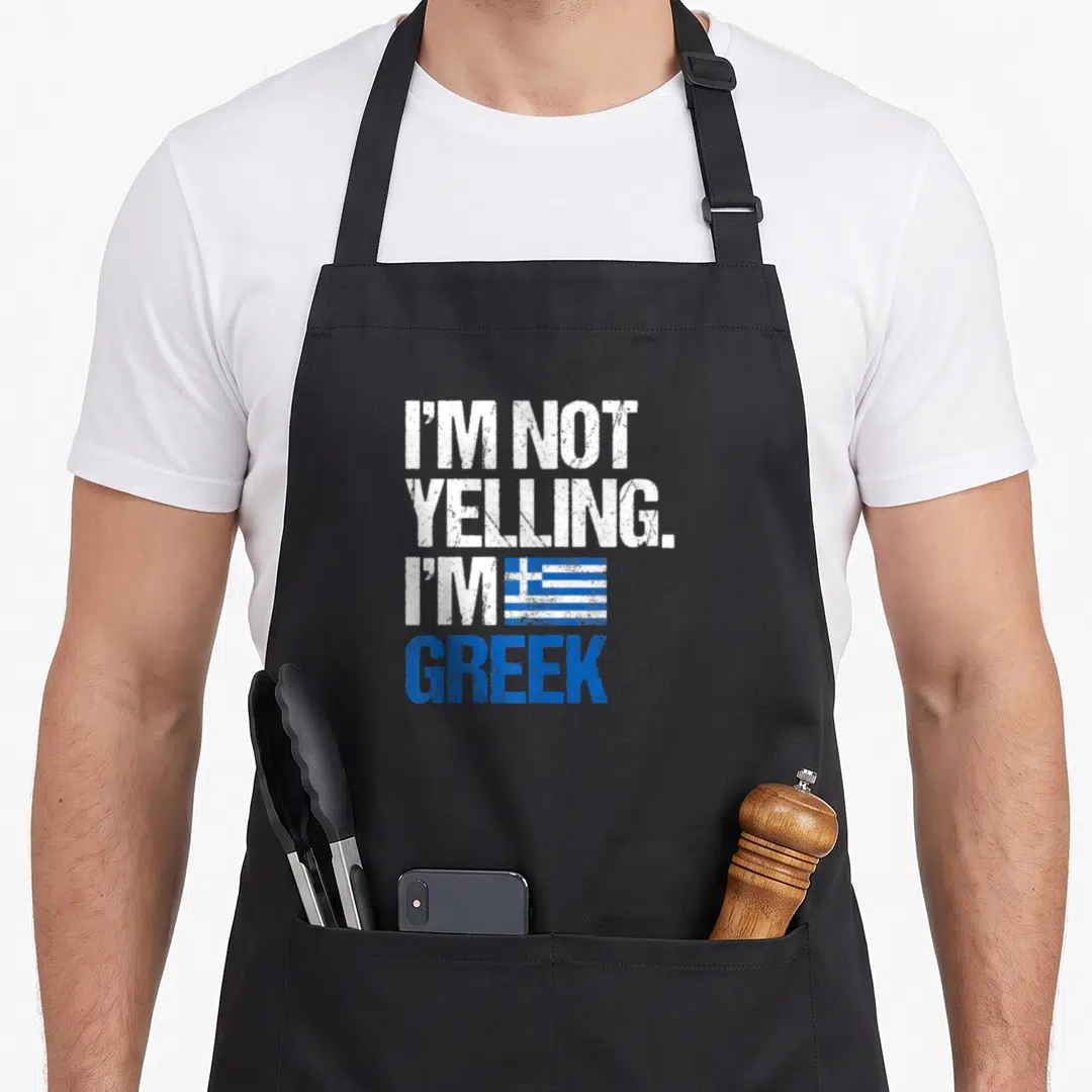Yelling greek aprons - Stacked white and blue distressed text reading I'M NOT YELLING I'M GREEK with a small flag — shown on a black cotton canvas apron against a white background