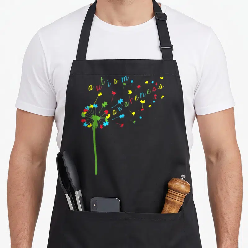Funny autism ribbon apron - Dandelion made of red blue yellow and green puzzle pieces forming autism awareness text — shown on a black cotton canvas apron against a white background