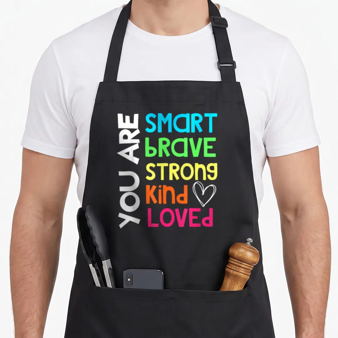 Smart teacher apron - Colorful text reading You Are Smart Brave Strong Kind Loved with a heart outline — shown on a black cotton canvas apron against a white background