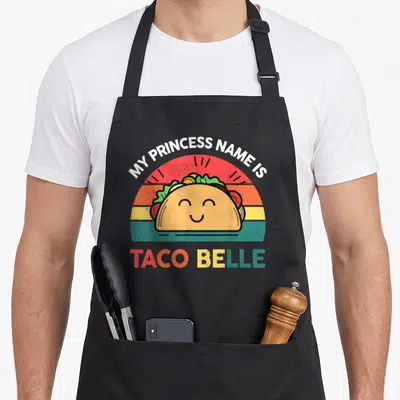 Taco princess belle apron - Smiling taco centered under arched text and above colorful block letters on a striped — shown on a black cotton canvas apron against a white background