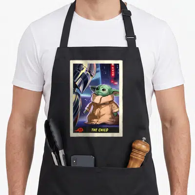 Trading card apron - Grogu standing next to a silver helmet under the text THE CHILD in a beige card frame — shown on a black cotton canvas apron against a white background