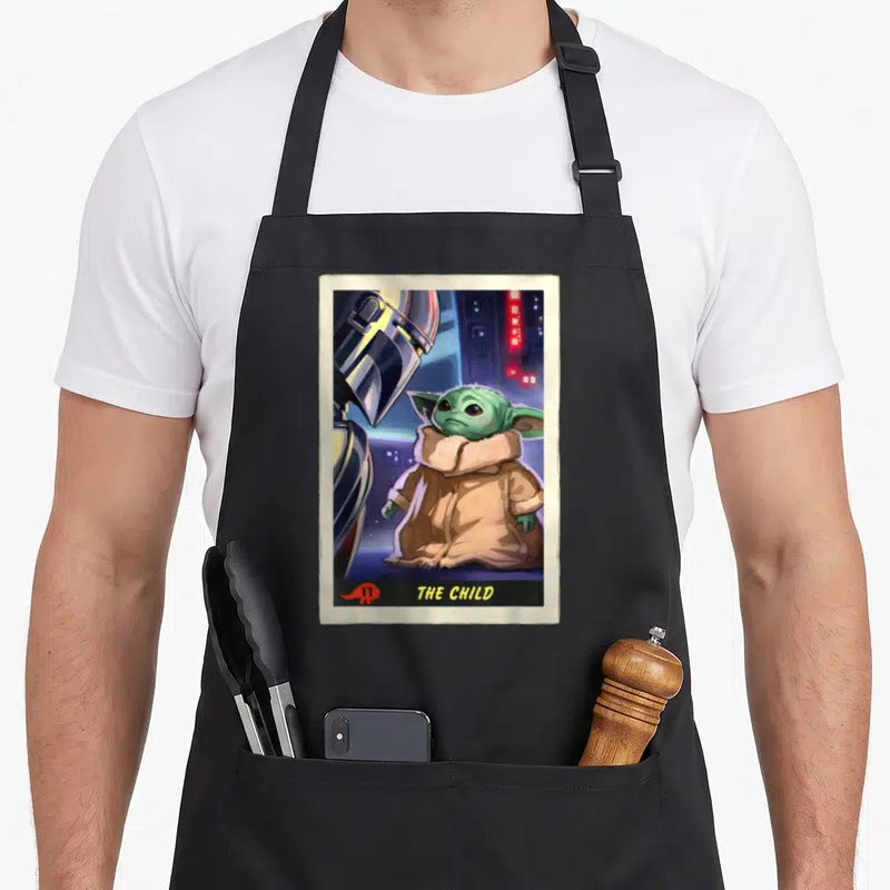 Trading card apron - Grogu standing next to a silver helmet under the text THE CHILD in a beige card frame — shown on a black cotton canvas apron against a white background