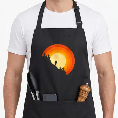 Backpacking hiking camping apron - Minimalist hiker silhouette on grey slope against striped orange and yellow sun — shown on a black cotton canvas apron against a white background