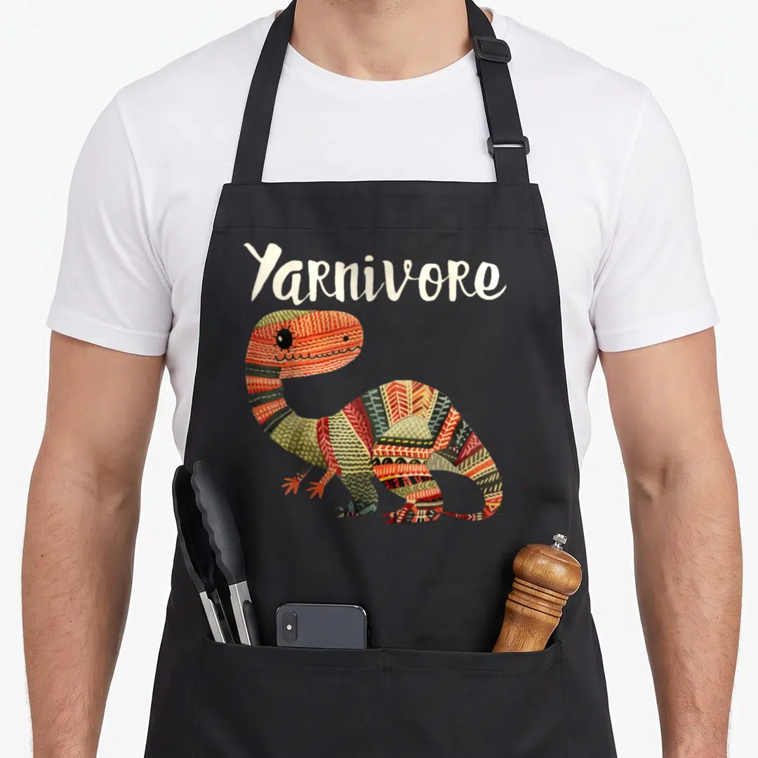 Yarnivore knitting apron - White Yarnivore text above a patterned dinosaur with knit-style body segments — shown on a black cotton canvas apron against a white background