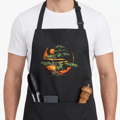 Buddhist tree apron - Cartoon green bonsai tree over orange circular sunset background — shown on a black cotton canvas apron against a white background