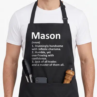 Definition mason apron - White text on grey background defining Mason as handsome, humble, and a master of trades — shown on a black cotton canvas apron against a white background
