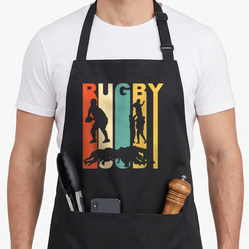 Funny rugby 1970s apron - Rugby text with grey player silhouettes on red, beige, blue, yellow, and grey vertical stripes — shown on a black cotton canvas apron against a white background