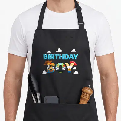 Cloud camping apron - BIRTHDAY BOY text formed by cowboy and space gear with five white clouds — shown on a black cotton canvas apron against a white background