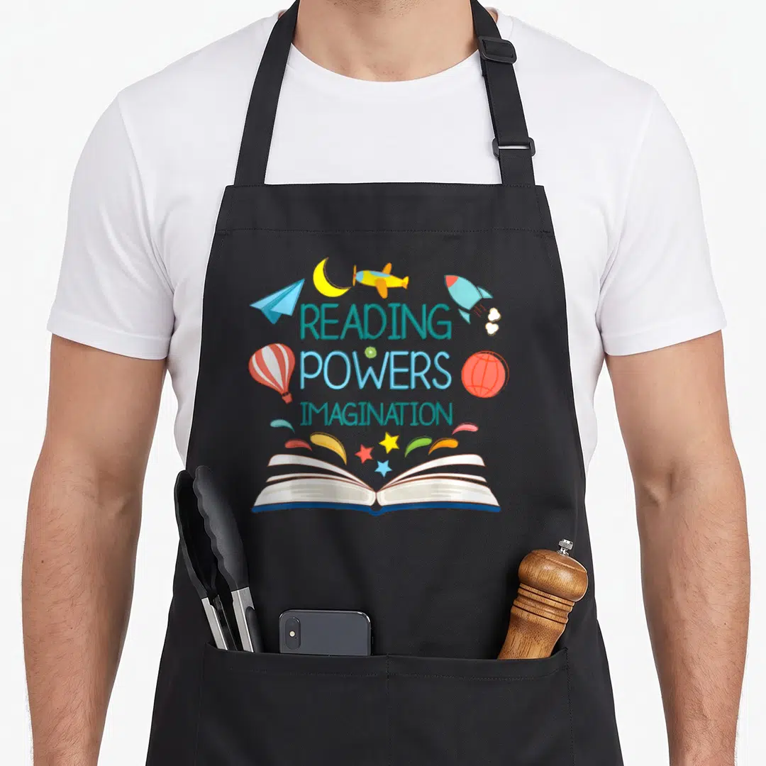 Power teacher apron - Open book with plane rocket balloon and stars surrounding stacked text reading powers imagination — shown on a black cotton canvas apron against a white background