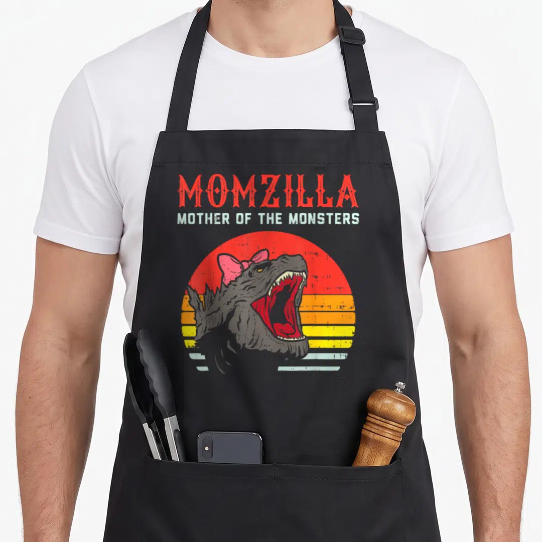 Momzilla mother's apron - MOMZILLA text above grey monster head with pink bow against red and yellow retro sunset — shown on a black cotton canvas apron against a white background
