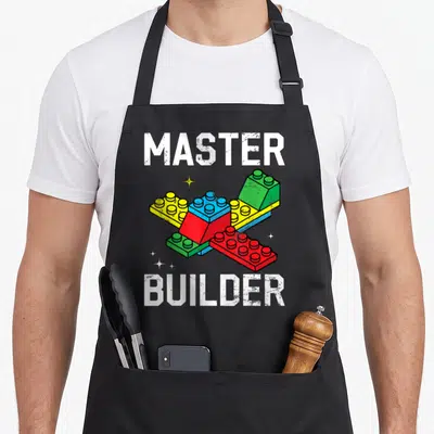 Block brick building apron - Distressed MASTER BUILDER text above red blue yellow green and white interlocking toy — shown on a black cotton canvas apron against a white background