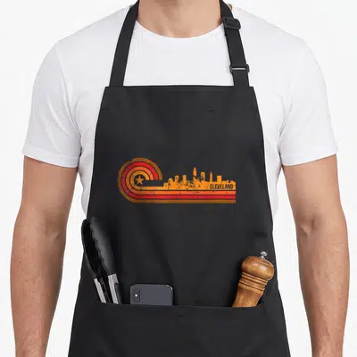 Cleveland skyline art apron - Retro orange and red skyline graphic with concentric circle star motif and distressed — shown on a black cotton canvas apron against a white background
