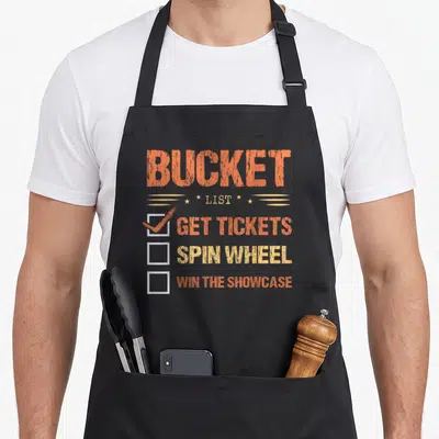 Showcase bucket apron - Distressed orange text BUCKET LIST GET TICKETS SPIN WHEEL WIN THE SHOWCASE with three checkboxes — shown on a black cotton canvas apron against a white background