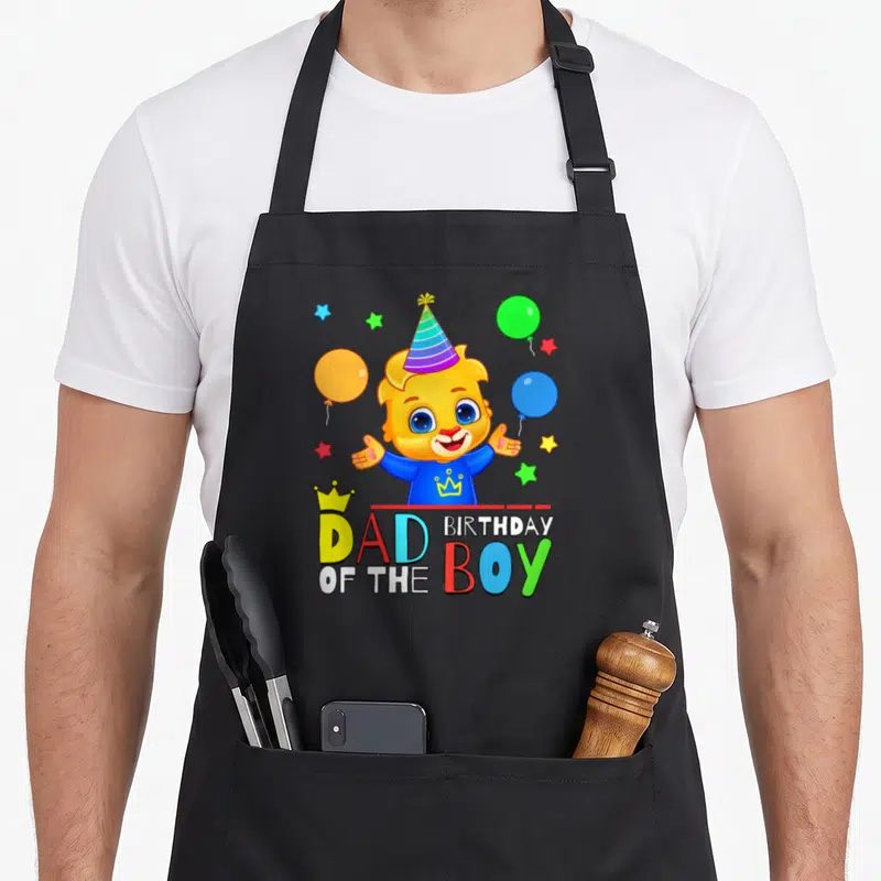 Celebrate lucas birthday apron - Cartoon lion cub with party hat and balloons above Dad of the Birthday Boy text — shown on a black cotton canvas apron against a white background