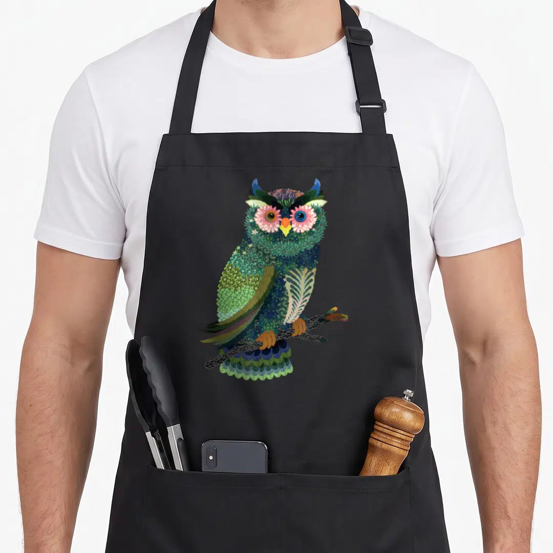 Owl floral apron - Cartoon owl made of green blue purple and pink floral patterns perched on a dark branch — shown on a black cotton canvas apron against a white background