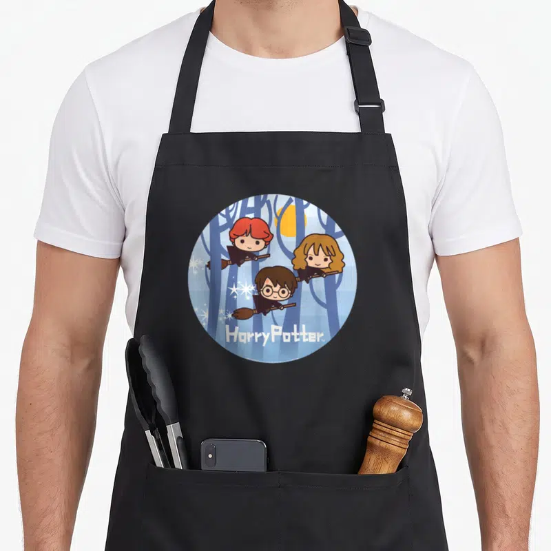 Chibi flying apron - Three cartoon characters flying on broomsticks in blue forest with yellow moon and Harry Potter — shown on a black cotton canvas apron against a white background