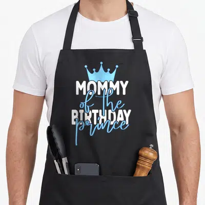 Mum prince birthday apron - Blue crown above white and blue MOMMY of the BIRTHDAY prince typography — shown on a black cotton canvas apron against a white background
