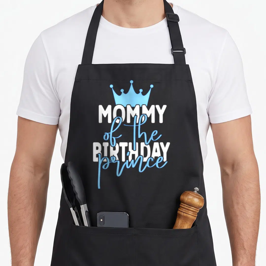 Mum prince birthday apron - Blue crown above white and blue MOMMY of the BIRTHDAY prince typography — shown on a black cotton canvas apron against a white background