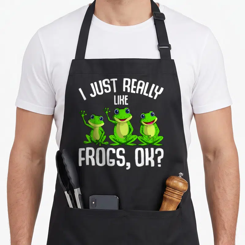 Frog aprons with pockets - Three green cartoon frogs lined up with white text I JUST REALLY LIKE FROGS OK — shown on a black cotton canvas apron against a white background
