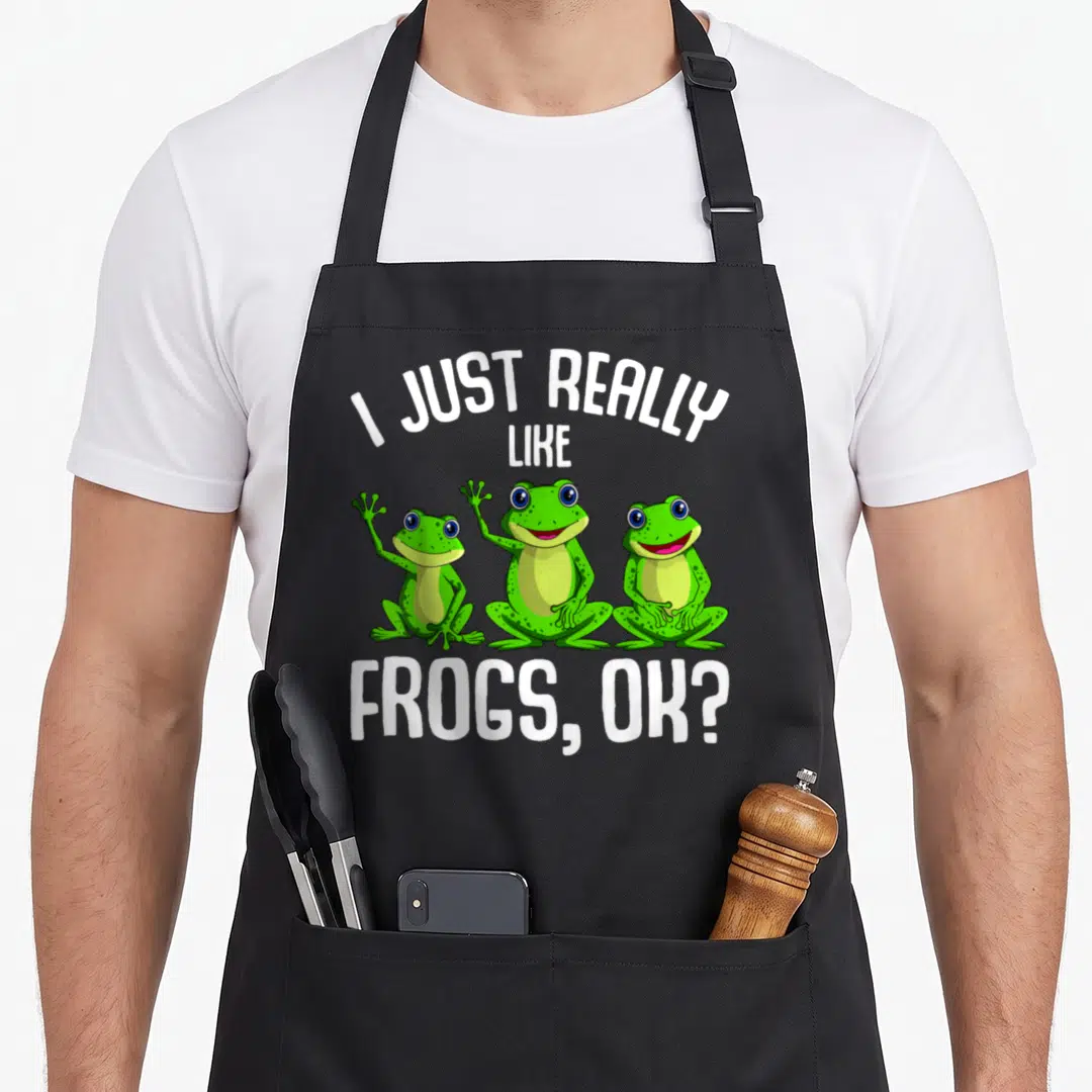 Frog aprons with pockets - Three green cartoon frogs lined up with white text I JUST REALLY LIKE FROGS OK — shown on a black cotton canvas apron against a white background
