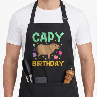 Capy birthday apron - Cartoon brown capybara between mint CAPY and yellow BIRTHDAY text with party items — shown on a black cotton canvas apron against a white background