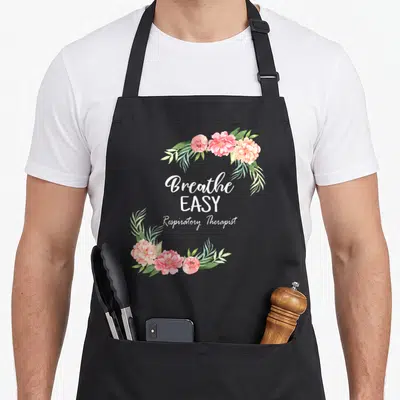 Respiratory floral apron - Watercolor floral frame with Breathe EASY Respiratory Therapist text in white script — shown on a black cotton canvas apron against a white background