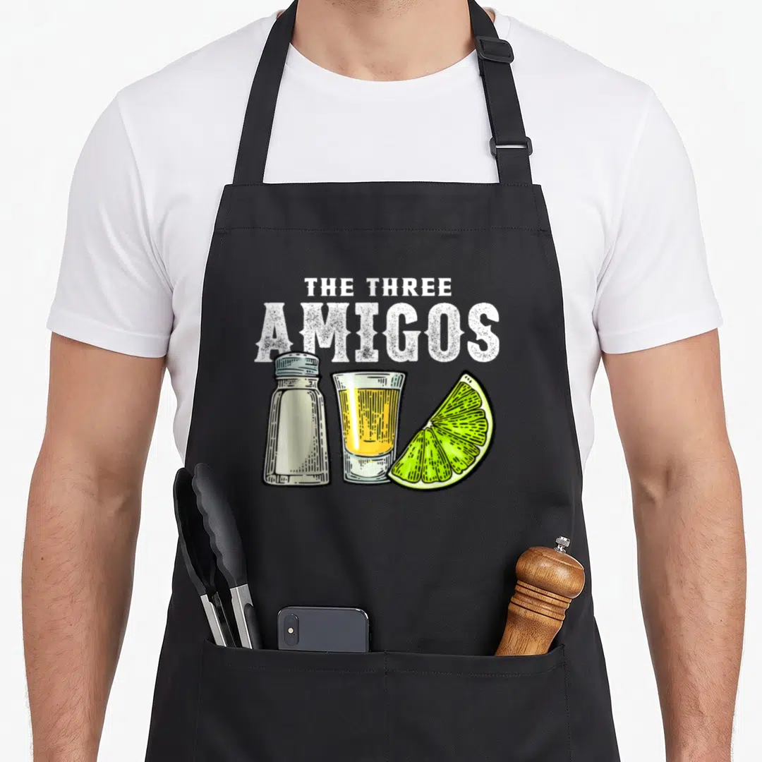 Three mayo apron - The Three Amigos text above salt shaker, tequila shot glass, and lime wedge in cartoon style — shown on a black cotton canvas apron against a white background