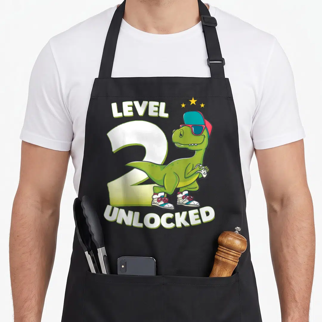 2nd birthday t-rex apron - Green cartoon dinosaur holding game controller in front of white number 2 with text — shown on a black cotton canvas apron against a white background