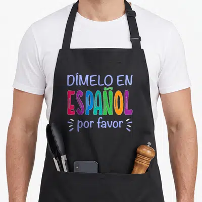 Dimelo spanish aprons - Stacked colorful typography reading DÍMELO EN ESPAÑOL por please in blue pink purple teal green — shown on a black cotton canvas apron against a white background