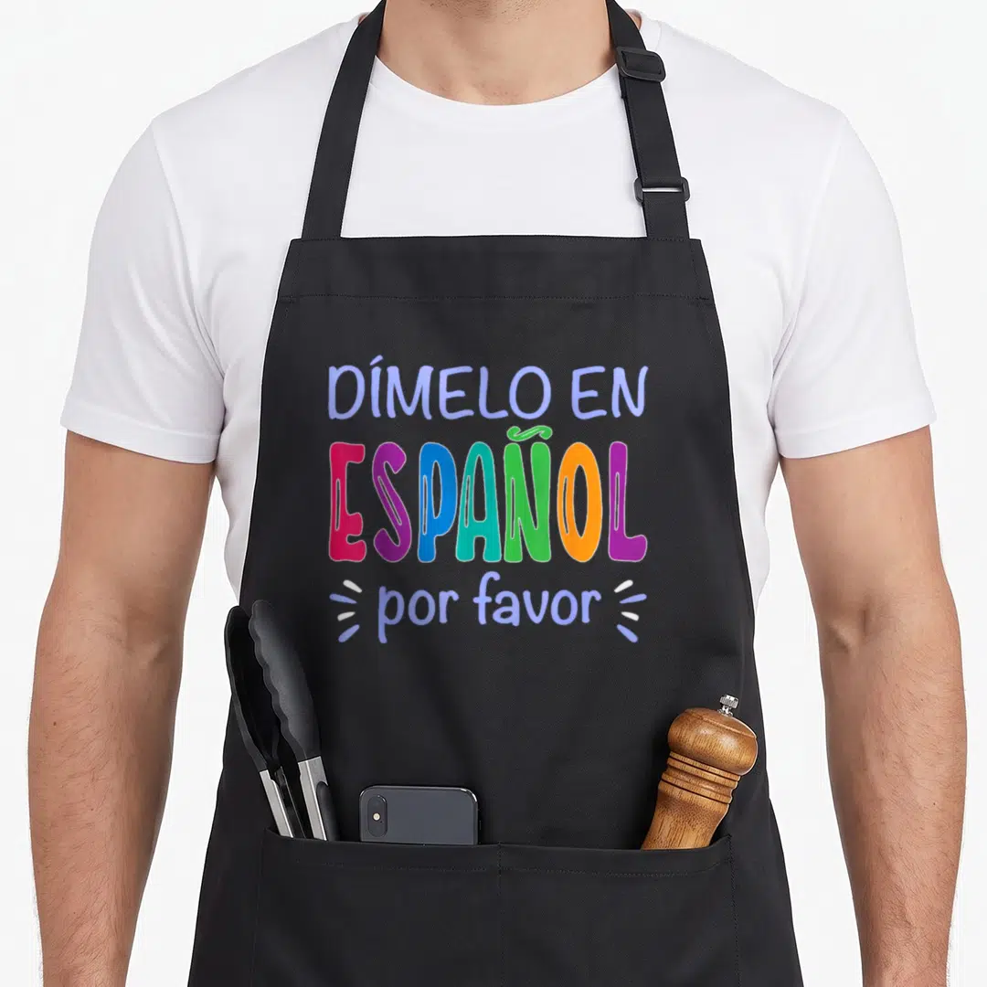 Dimelo spanish aprons - Stacked colorful typography reading DÍMELO EN ESPAÑOL por please in blue pink purple teal green — shown on a black cotton canvas apron against a white background