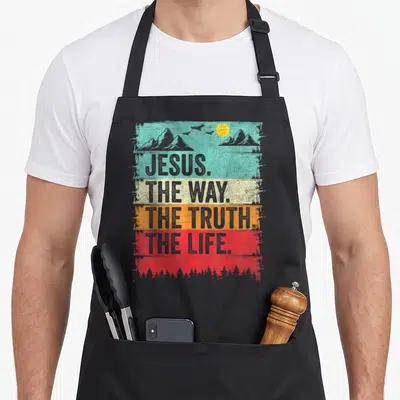 Way christian apron - Four colored bands reading JESUS THE WAY THE TRUTH THE LIFE over mountains sun birds and tree — shown on a black cotton canvas apron against a white background
