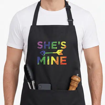 Couple she's in her apron - SHE'S MINE rainbow gradient text with heart arrow on black bib apron — shown on a black cotton canvas apron against a white background