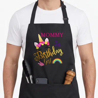 Cute birthday unicorn apron - MOMMY OF THE Birthday Girl text with unicorn head, flowers, rainbow, and pink wing — shown on a black cotton canvas apron against a white background