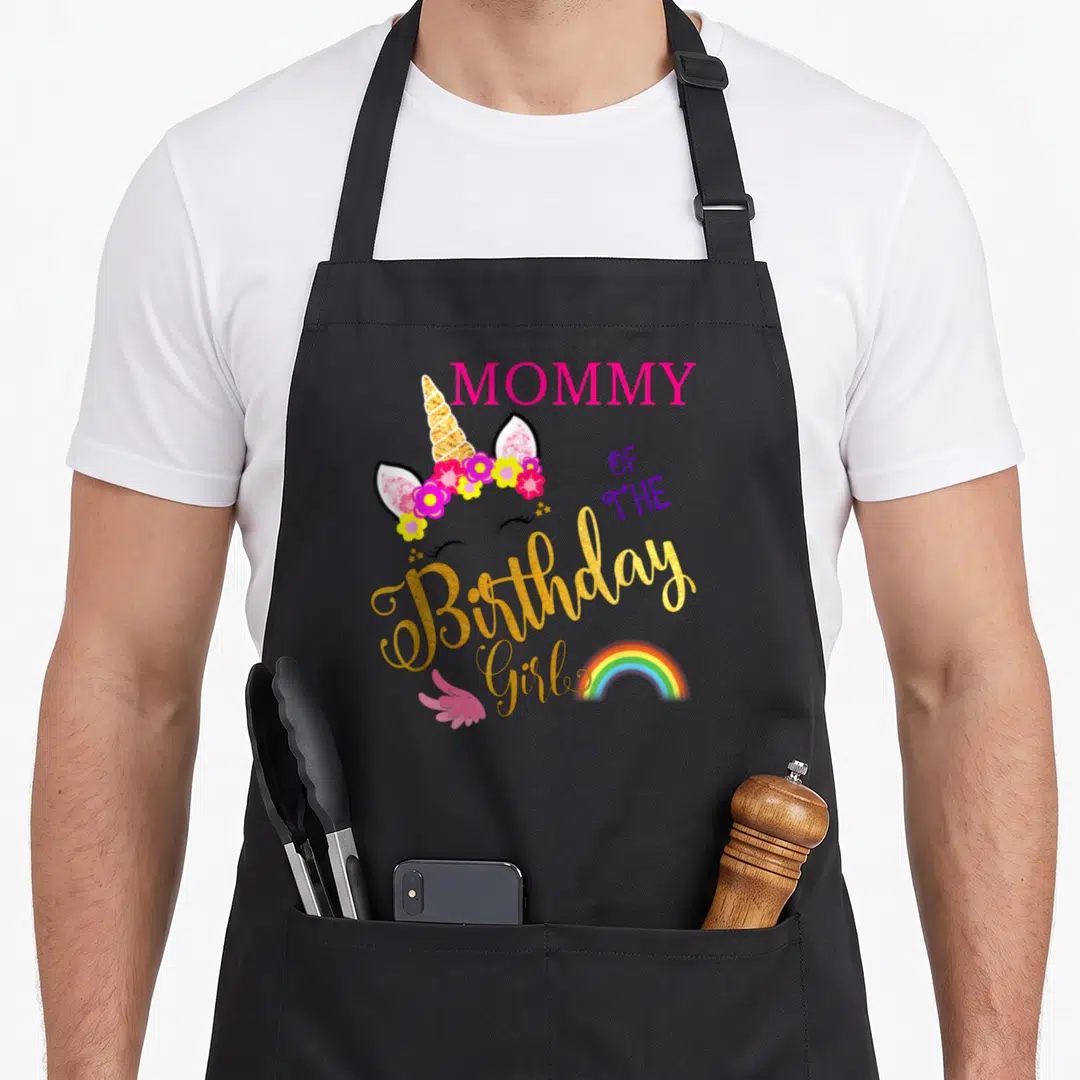 Cute birthday unicorn apron - MOMMY OF THE Birthday Girl text with unicorn head, flowers, rainbow, and pink wing — shown on a black cotton canvas apron against a white background