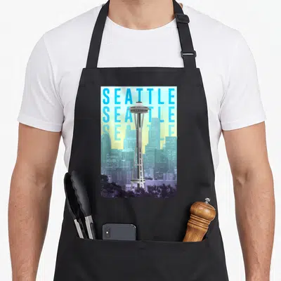 Space seattle apron - Space Needle tower overlapping three stacked SEATTLE text lines with grey purple skyline and trees — shown on a black cotton canvas apron against a white background