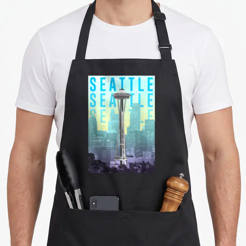 Space seattle apron - Space Needle tower overlapping three stacked SEATTLE text lines with grey purple skyline and trees — shown on a black cotton canvas apron against a white background