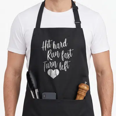 Turn baseball apron - White distressed text Hit hard Run fast Turn left above a heart-shaped baseball with stitching — shown on a black cotton canvas apron against a white background
