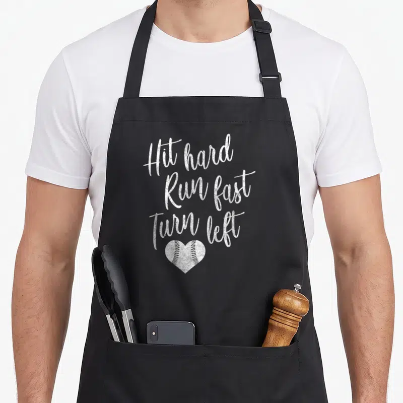 Turn baseball apron - White distressed text Hit hard Run fast Turn left above a heart-shaped baseball with stitching — shown on a black cotton canvas apron against a white background