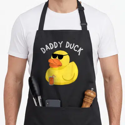 Daddy rubber cotton duck apron - Yellow rubber duck with sunglasses and orange drink under arched DADDY DUCK text — shown on a black cotton canvas apron against a white background