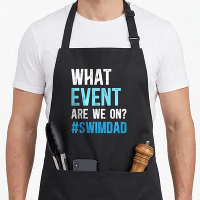 Father swimmer swimming dad apron - White and blue stacked text reading WHAT EVENT ARE WE ON? #SWIMDAD — shown on a black cotton canvas apron against a white background