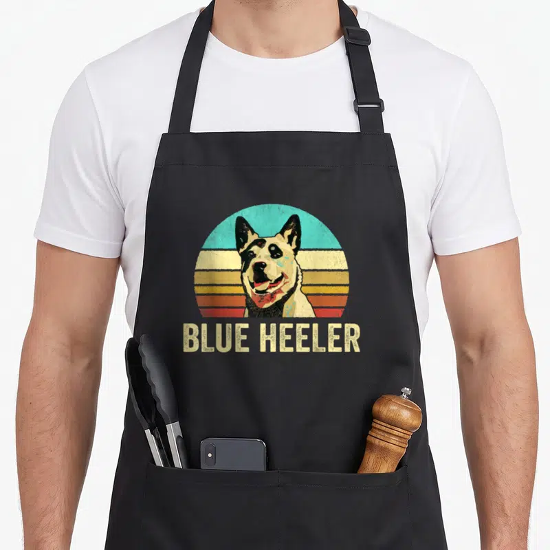 Sunset dog apron - Retro Blue Heeler head with striped sunset and BLUE HEELER text — shown on a black cotton canvas apron against a white background