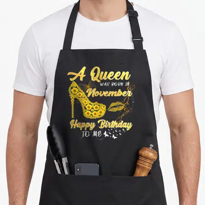 November sunflower apron - Gold text A Queen Was Born In November around sunflower high heel shoe and butterfly graphics — shown on a black cotton canvas apron against a white background