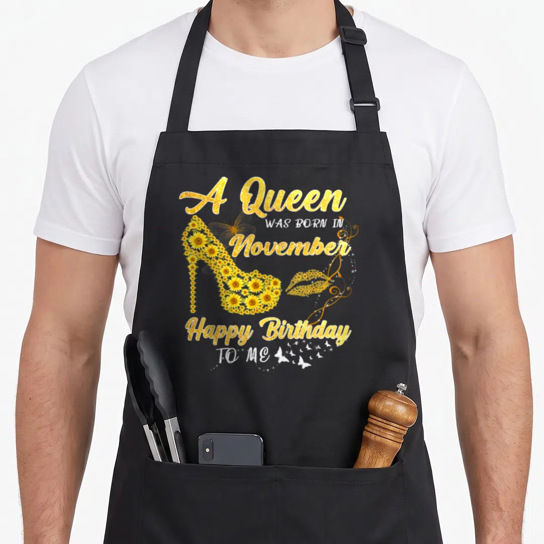 November sunflower apron - Gold text A Queen Was Born In November around sunflower high heel shoe and butterfly graphics — shown on a black cotton canvas apron against a white background