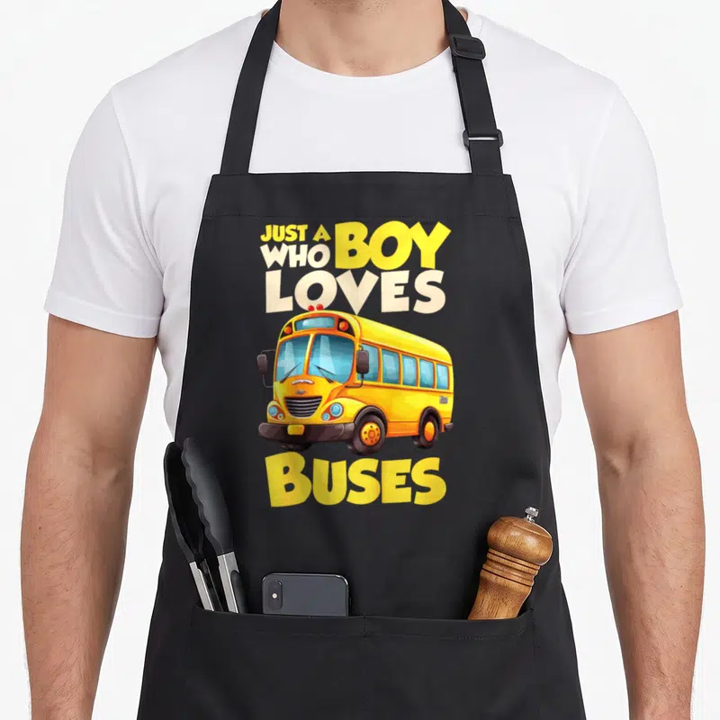 Funny buse school bus apron - Cartoon yellow school bus with text JUST A BOY WHO LOVES BUSES in white and blue lettering — shown on a black cotton canvas apron against a white background