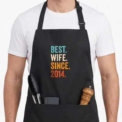 Wife wedding apron - Stacked BEST WIFE SINCE 2014 text in blue beige orange and red distressed letters — shown on a black cotton canvas apron against a white background