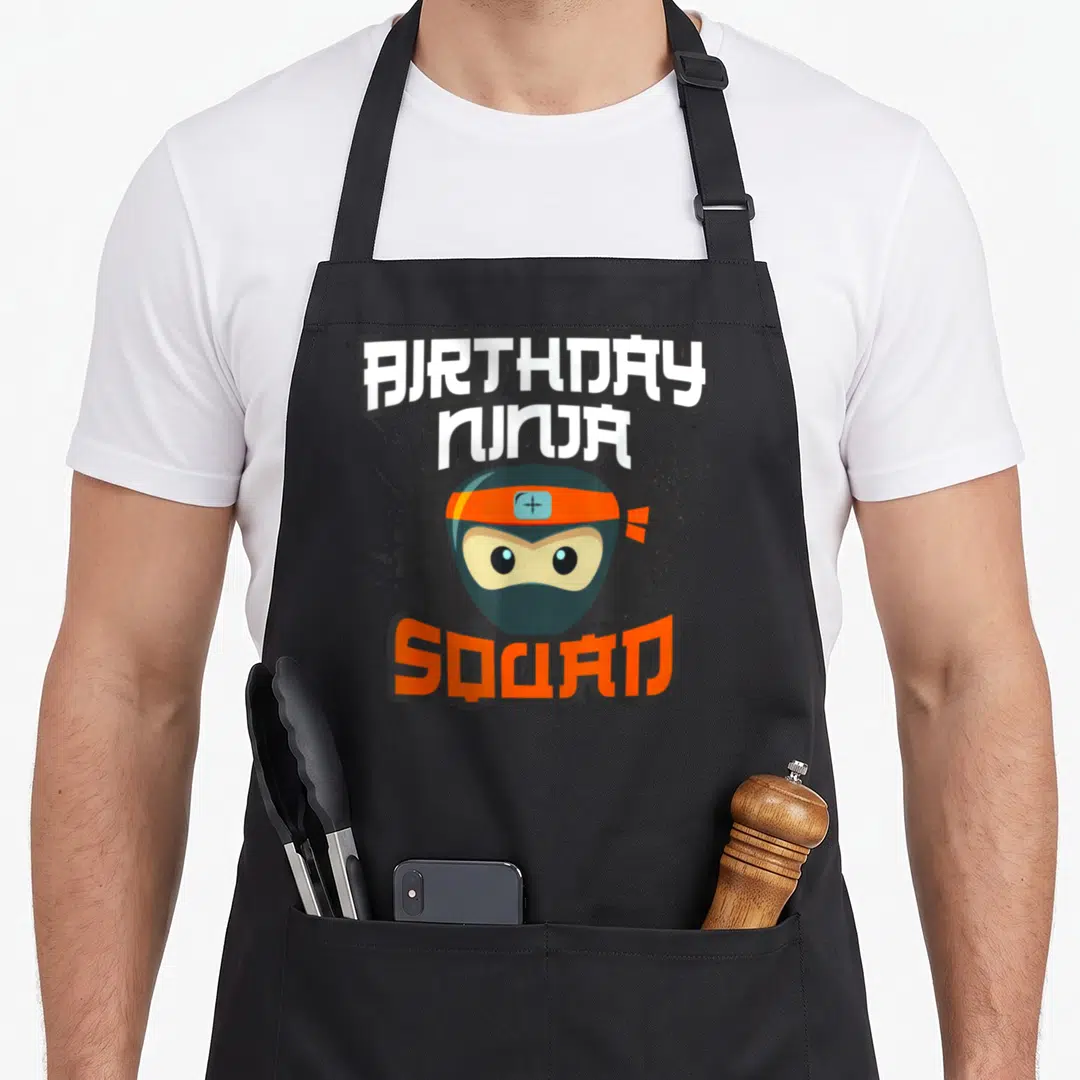 Ninja squad birthday apron - Cartoon ninja head with orange headband and BIRTHDAY NINJA SQUAD text on grey splatter — shown on a black cotton canvas apron against a white background