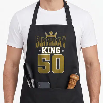 Theme kings apron - Gold and white BIRTHDAY KING 50 text with crown and stars on black bib — shown on a black cotton canvas apron against a white background