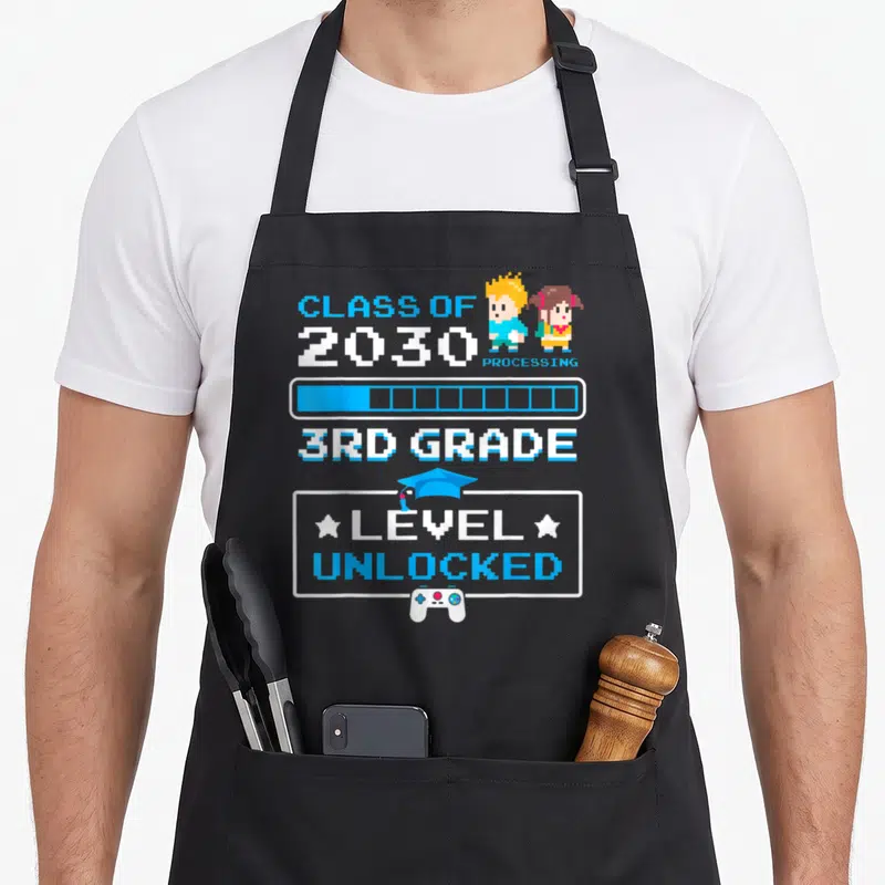 3rd grade video game apron - Pixel art text Class of 2030 Processing 3rd Grade Level Unlocked with characters — shown on a black cotton canvas apron against a white background